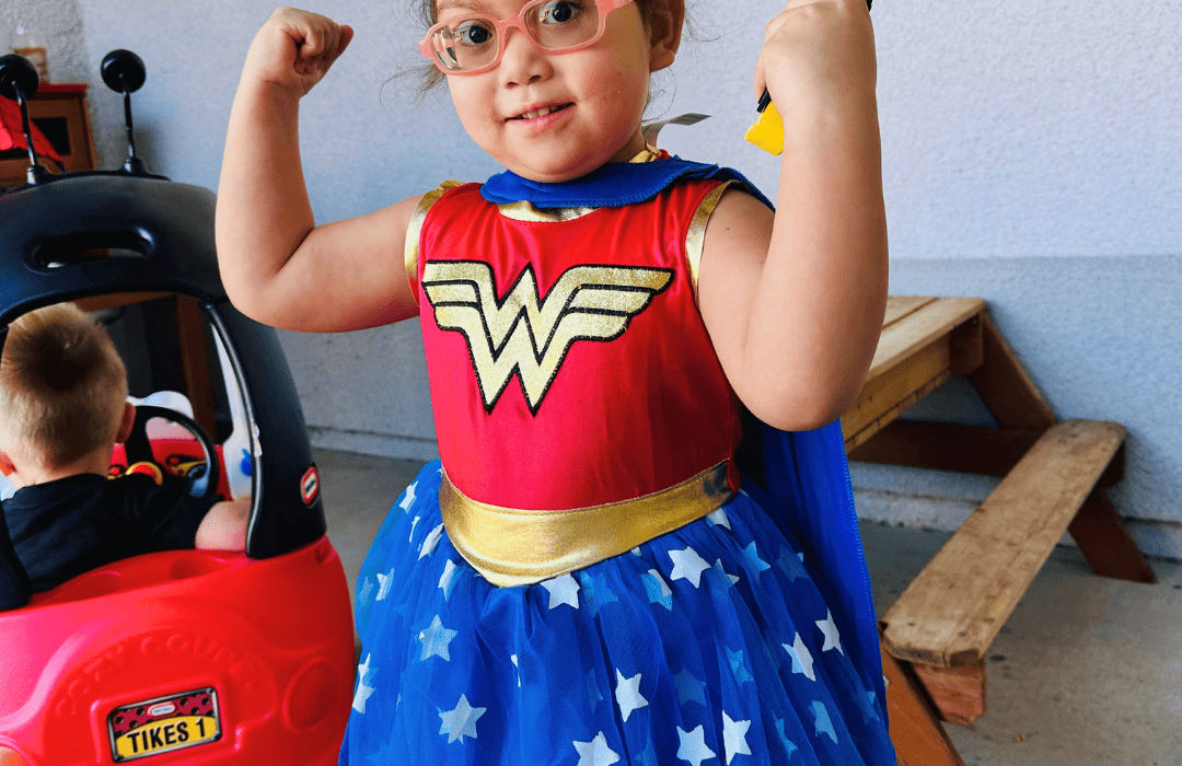 A little girl wearing a Wonder Woman costume gives a power pose and smiles for the camera.