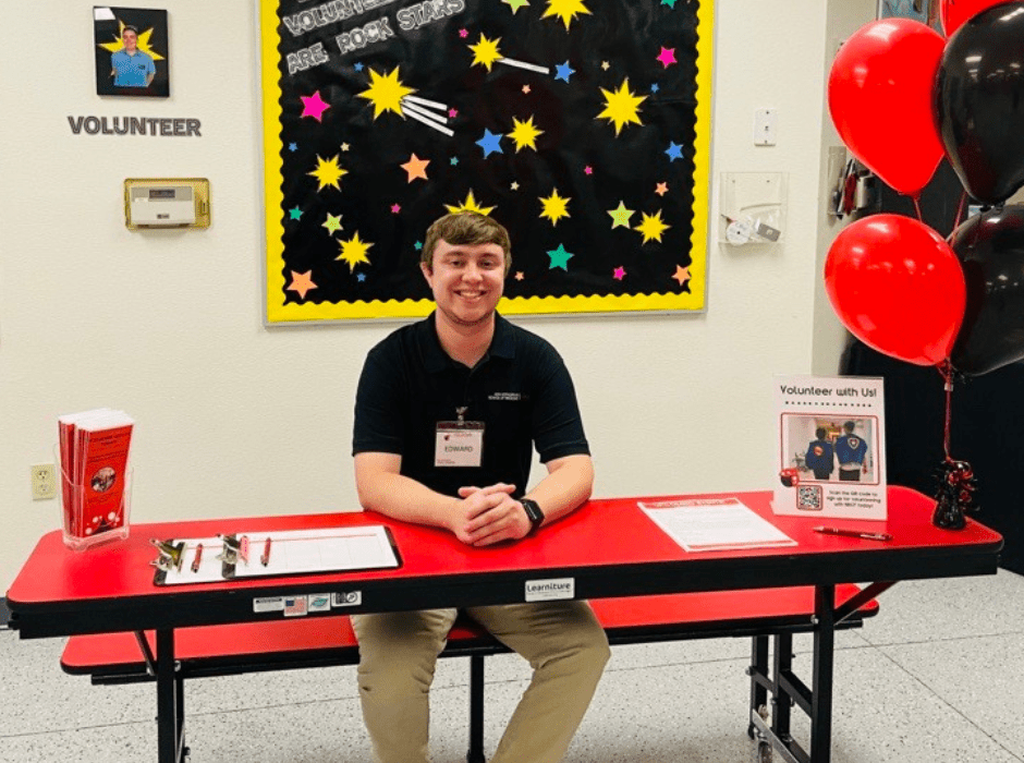 NBCF volunteer, Ed Pizio, sits at the volunteer table at NBCF's Open House. Behind him is a volunteer board that reads, "Our Volunteers Are Rock Stars!" and is decorated with various stars. The table has pamphlets on how to volunteer and red and black balloons.