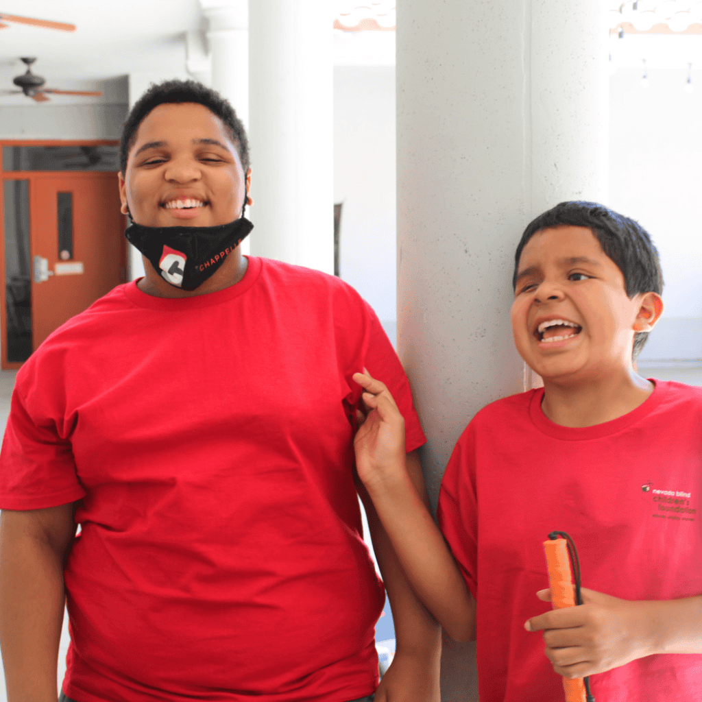 Image shows Alexis laughing while standing next to his smiling best friend, Amir, while they stand in the sunny courtyard of NBCF. Both boys wear red NBCF shirts.
