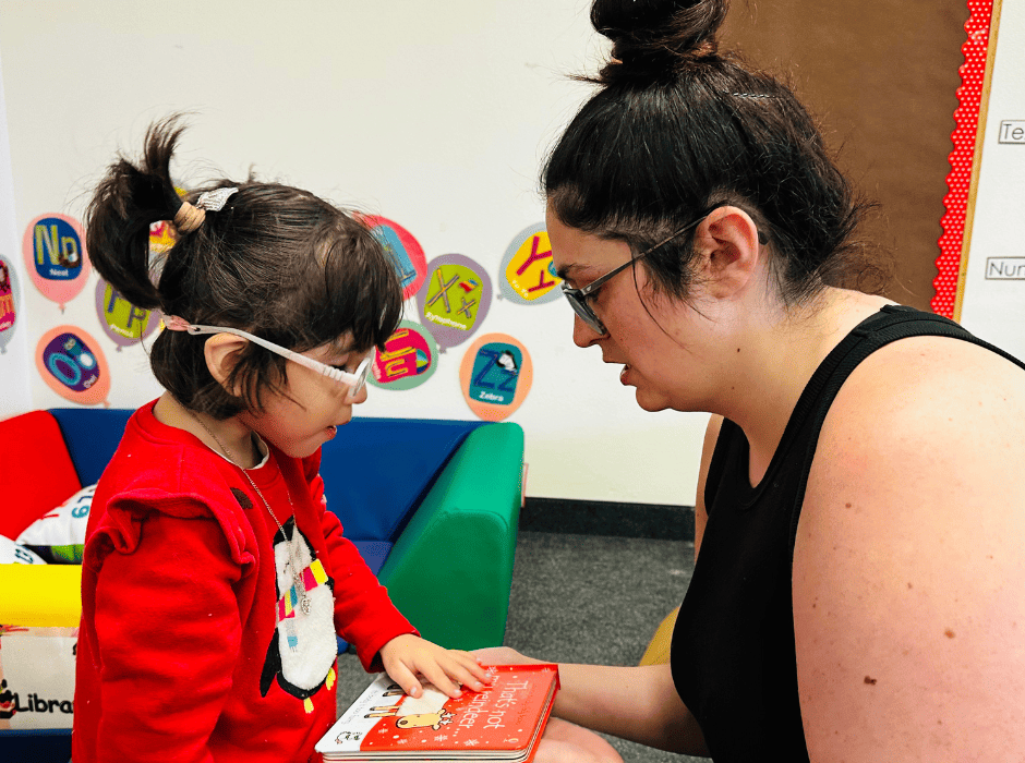Miss Megan kneels in front of a little girl wearing pink glasses. She shows her a children's braille book.